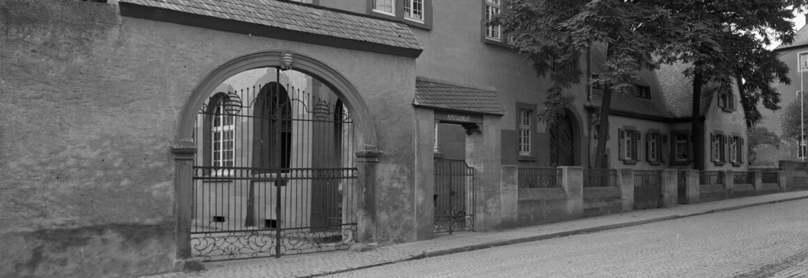 Ein Gebäude mit einem großen, verzierten Torbogen aus Metall, das in den Innenhof vom Domänenhof führt. Rechts daneben steht befindet sich der Eingang zum Hochheimer "Amtsgericht".