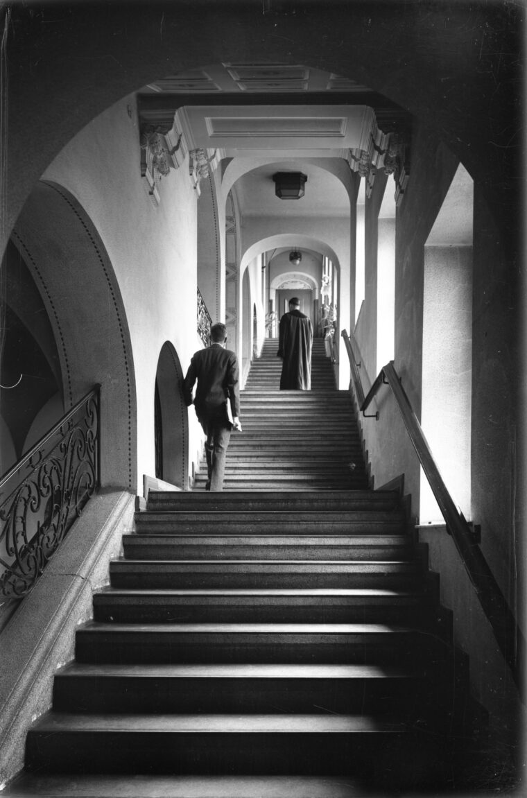 Eine Innenaufnahme eines Treppenhauses vom Amtsgericht, in dem zwei Personen eine lange Treppe hinaufsteigen. Der Flur ist von Bögen und kunstvollen Geländern gesäumt und wirkt groß und imposant.