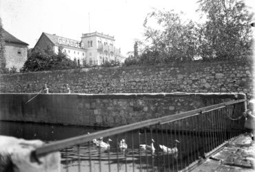 Das Schwarz-Weiß-Foto zeigt die Hochheimer Backeswied mit der Graeger Villa im Hintergrund. Im Vordergrund ist ein Wassergraben zu sehen, in dem mehrere Enten schwimmen. Eine Steinmauer verläuft entlang des Grabens. Zwei Kinder sind auf der Mauer zu sehen, während ein weiteres Kind rechts an einem Geländer steht. Im Hintergrund erhebt sich die markante, mehrstöckige Villa mit der Aufschrift 'GRAEGER' auf dem Dach.