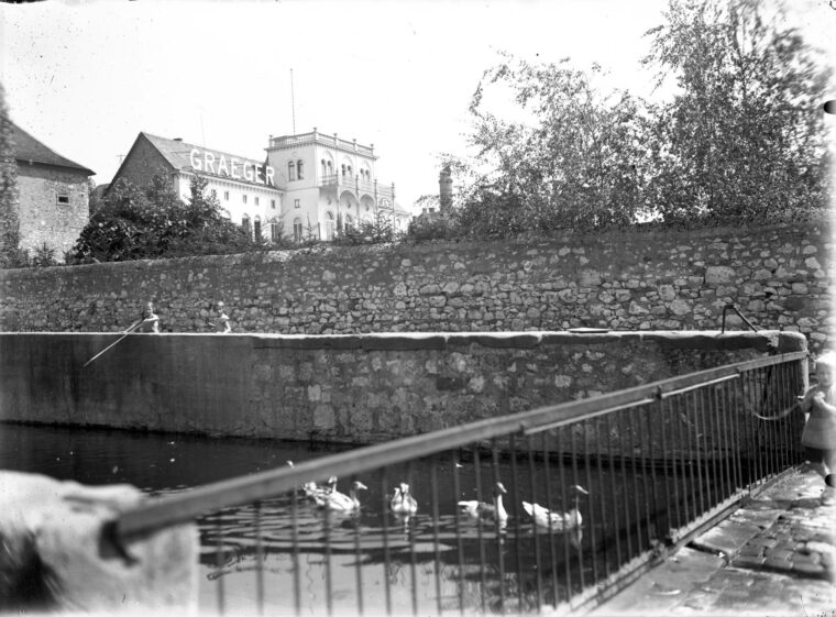 Das Schwarz-Weiß-Foto zeigt die Hochheimer Backeswied mit der Graeger Villa im Hintergrund. Im Vordergrund ist ein Wassergraben zu sehen, in dem mehrere Enten schwimmen. Eine Steinmauer verläuft entlang des Grabens. Zwei Kinder sind auf der Mauer zu sehen, während ein weiteres Kind rechts an einem Geländer steht. Im Hintergrund erhebt sich die markante, mehrstöckige Villa mit der Aufschrift 'GRAEGER' auf dem Dach.