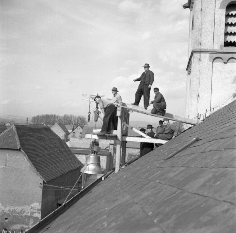 Das Schwarzweißfoto zeigt mehrere Männer, die auf einem improvisierten Holzgerüst arbeiten, das an einem Kirchturm angebracht ist. Sie scheinen dabei zu sein, eine große Glocke mithilfe eines Flaschenzugs in den Kirchturm zu heben. Im Hintergrund sind Dächer und Häuser zu sehen.