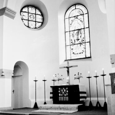 Das Schwarzweißfoto zeigt das Innere der evangelischen Kirche in Hochheim. Im Vordergrund befindet sich der Altar, der mit Blumen und Kerzen geschmückt ist, dahinter ein großes Kruzifix. An der Wand hinter dem Altar sind zwei bunte Glasfenster zu sehen, die von außen einfallendes Licht hereinlassen.