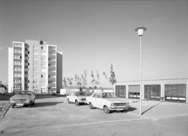 Ein schwarz-weißes Foto zeigt einen Parkplatz mit mehreren Autos vor einem modernen Hochhaus. Im Hintergrund ist ein weiteres, niedrigeres Gebäude zu sehen.