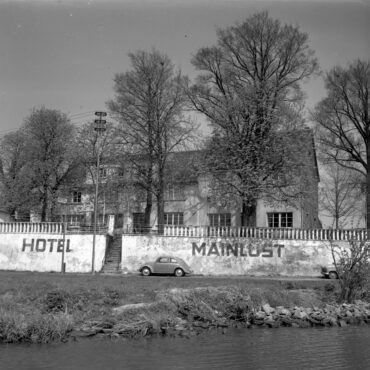 Das Foto zeigt das Hotel Mainlust, das hinter einer weißen Mauer mit großen Bäumen im Hintergrund liegt. Zwei Autos parken vor dem Hotel, und das Gebäude ist von einer ruhigen, ländlichen Atmosphäre umgeben.