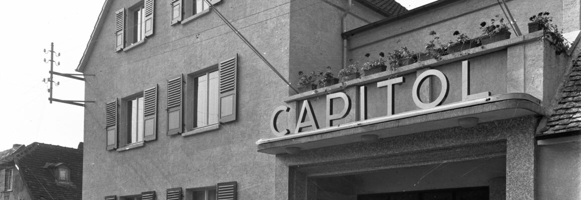 Ein schlichtes Gebäude mit einem Vordach und der Aufschrift "Capitol" über dem Eingang. Die Fensterläden des Hauses sind geöffnet, und auf dem Dach blühen Pflanzen in Kästen.