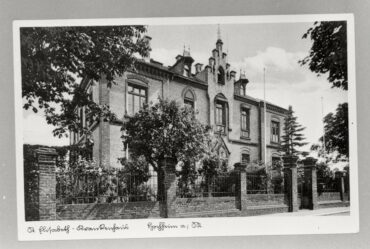 Das Foto zeigt das Gebäude vom St. Elisabeth Krankenhaus. Es hat eine aufwendig gestaltete Ziegelfassade, die umgeben ist von einem gepflegten Garten mit Bäumen und einem eisernen Zaun. Das Bild zeigt die Vorderansicht des Anwesens bei sonnigem Wetter.