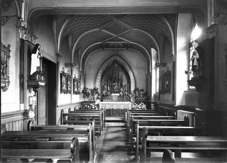 Das Foto in Schwarzweiß zeigt die Krankenhaus-Kapelle mit gotischem Gewölbe und einem reich verzierten Altar, flankiert von Statuen und Bänken aus Holz. Die Wände sind mit religiösen Darstellungen und Blumenarrangements geschmückt.