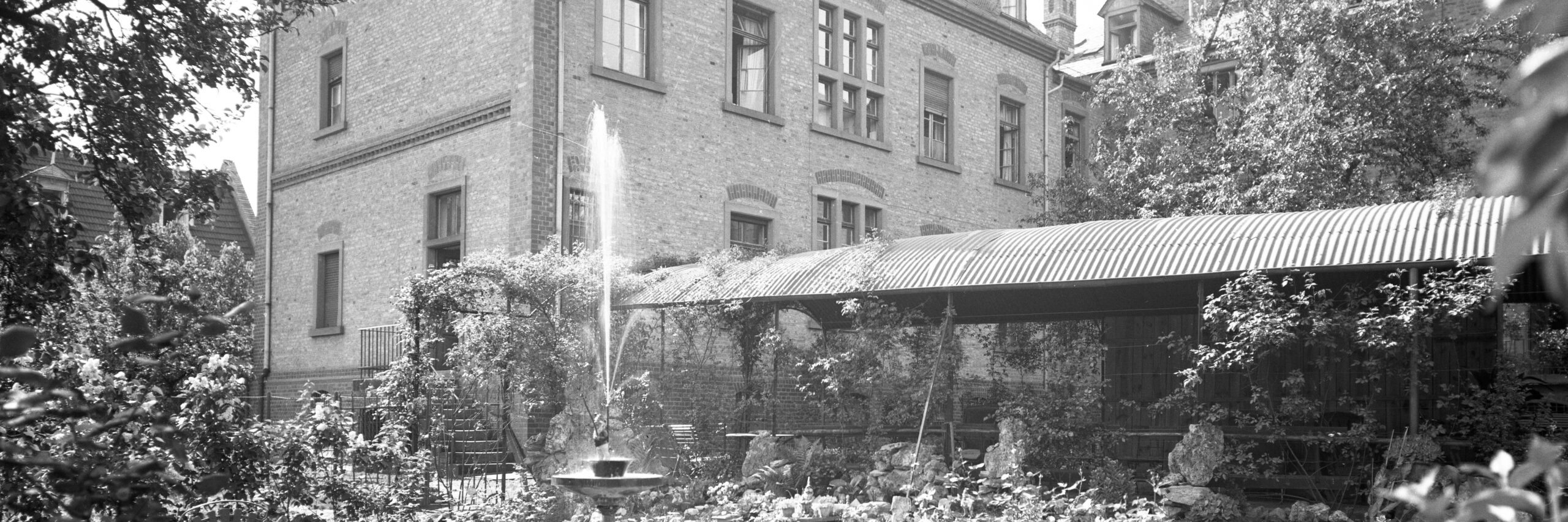 Das Foto zeigt den Garten des Krankenhauses mit einem Brunnen und Parkbänken vor einem Backsteingebäude. Der Garten ist üppig bepflanzt und bietet einen ruhigen Rückzugsort für Patienten und Besucher.