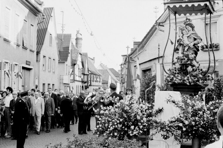 Eine Prozession von Menschen, die an der reich geschmückten Madonna am Plan stehen. Die Straße ist mit Blumen und Flaggen dekoriert, und die Häuser im Hintergrund sind mit Festtagsdekorationen geschmückt.