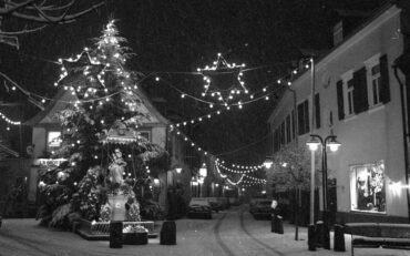 Die Madonna am Plan ist festlich beleuchtet und von weihnachtlicher Dekoration umgeben. Die Straße ist schneebedeckt, und Lichterketten hängen über der Straße, was eine gemütliche und festliche Stimmung erzeugt.