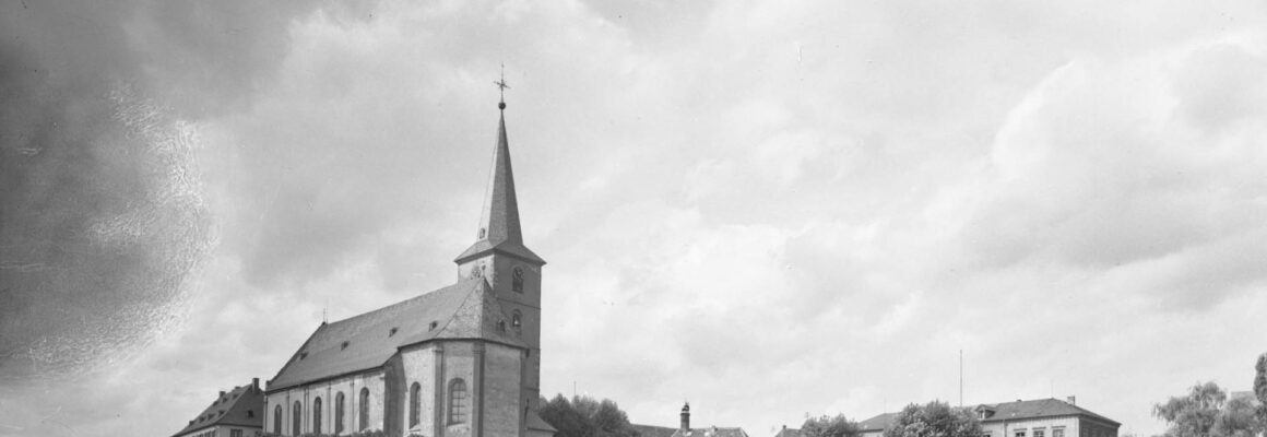 Die Pfarrkirche St. Peter und Paul ist von Weinbergen und einer ländlichen Straße umgeben. Die Kirche steht stolz auf einer kleinen Anhöhe, und der sonnige Himmel und die gepflegten Weinreben vermitteln eine friedliche und idyllische Atmosphäre.