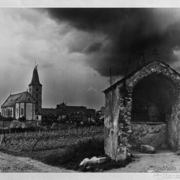 Ein Foto aus dem Jahr 1946 zeigt die zerstörte Pfarrkirche St. Peter und Paul im Hintergrund, deren Turm durch Kriegseinwirkungen beschädigt ist. Im Vordergrund ist eine kleine, verfallene Kapelle zu sehen, während dunkle Gewitterwolken den Himmel bedecken und eine bedrückende Atmosphäre schaffen.