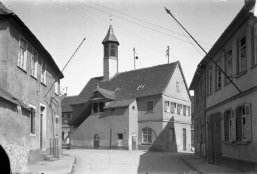 Das Schwarz-Weiß-Foto zeigt das alte Rathauses in Hochheim am Kälberplatz. Der Platz ist gepflastert und von alten Gebäuden umgeben. Im Mittelpunkt steht das Rathaus mit seinem auffälligen Turm und der Außentreppe. Elektrische Leitungen verlaufen über den Platz.