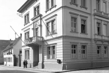 Das Schwarz-Weiß-Foto zeigt ein Gebäude, das als Rathaus in der Kirchstraße 13 diente. Es hat eine helle Fassade mit symmetrisch angeordneten Fenstern und einem dekorativen Eingang, der über eine kleine Treppe erreichbar ist. Zwei Personen stehen vor dem Eingang auf dem Kopfsteinpflaster der Straße. Im Hintergrund sind weitere ältere Gebäude zu sehen.