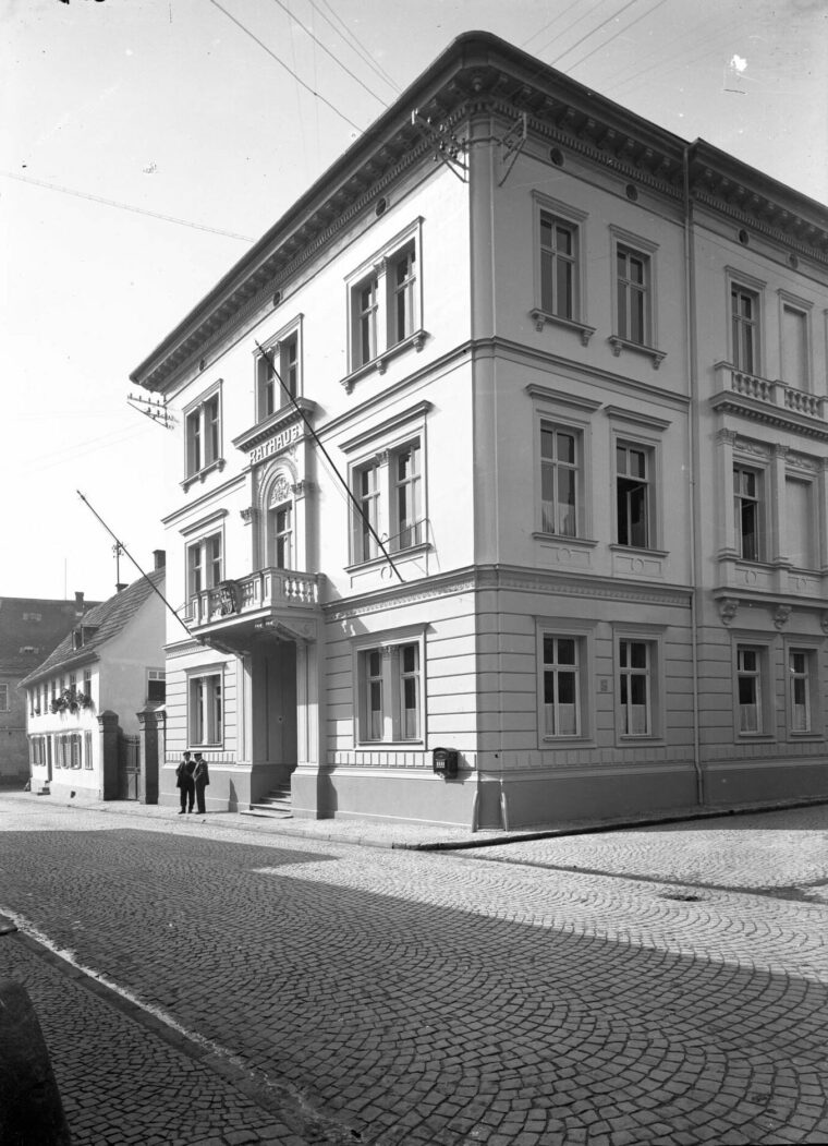 Das Schwarz-Weiß-Foto zeigt ein Gebäude, das als Rathaus in der Kirchstraße 13 diente. Es hat eine helle Fassade mit symmetrisch angeordneten Fenstern und einem dekorativen Eingang, der über eine kleine Treppe erreichbar ist. Zwei Personen stehen vor dem Eingang auf dem Kopfsteinpflaster der Straße. Im Hintergrund sind weitere ältere Gebäude zu sehen.