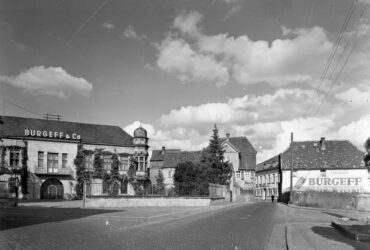 Das Foto zeigt ein Gebäude der Sektkellerei "Burgeff & Co." mit einem markanten Turm und zahlreichen Fenstern. Die Straße im Vordergrund ist gepflastert. Rechts ist eine große Werbetafel der Sektkellerei zu erkennen.