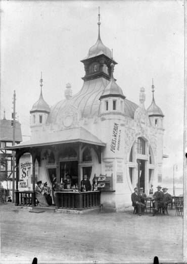 Der Schaumweinpavillon von Fuchs & Werum auf der Düsseldorfer Kunst- und Gewerbeausstellung zeigt ein prachtvolles Gebäude mit dekorativen Türmen. Vor dem Pavillon sitzen und stehen mehrere Personen, die Sekt trinken und sich unterhalten.