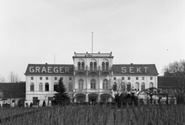 Die Fassade der Sektkellerei Carl Graeger, ein imposantes Gebäude mit der Aufschrift "Graeger Sekt" auf dem Dach. Vor dem Gebäude erstrecken sich Weinreben, die zu den Weinbergen der Kellerei gehören.