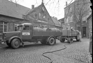 Ein Lastwagen, beladen mit großen Holzfässern, steht vor der Sektkellerei Carl Graeger. Im Hintergrund sind die historischen Gebäude der Kellerei zu sehen, und ein Arbeiter steht neben dem Lastwagen.