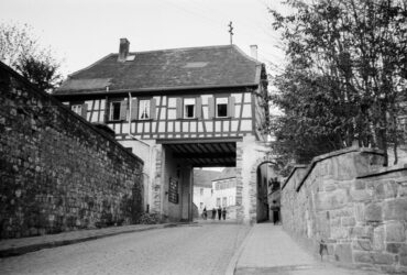 Das Foto zeigt das Hochheimer Stadttor mit einem Fachwerkgebäude darüber. Das Tor führt zu einer gepflasterten Straße, die von alten Steinmauern flankiert wird. Im Hintergrund sind mehrere Personen zu sehen, die die Straße entlanggehen.