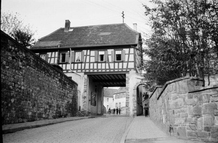 Das Foto zeigt das Hochheimer Stadttor mit einem Fachwerkgebäude darüber. Das Tor führt zu einer gepflasterten Straße, die von alten Steinmauern flankiert wird. Im Hintergrund sind mehrere Personen zu sehen, die die Straße entlanggehen.