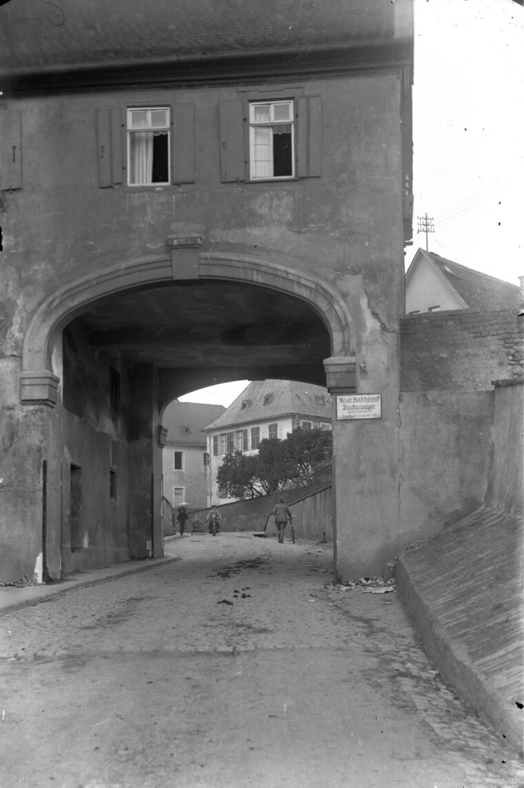 Das Bild zeigt das Hochheimer Stadttor mit einem darüberliegenden Fachwerkhaus, das zwei Fenster mit geöffneten Fensterläden hat. Unter dem Torbogen führt eine gepflasterte Straße weiter in die Ferne, auf der einige Menschen zu Fuß und mit Fahrrädern unterwegs sind.