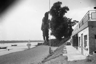 Das Bild zeigt das Hochheimer Strandbad am Ufer des Mains mit einem modernen Gebäude und einer Terrasse im Vordergrund. Am Sandstrand sind einige Boote und ein Segelboot auf dem Wasser zu sehen. Große Bäume säumen das Ufer und sorgen für eine ruhige und idyllische Atmosphäre.