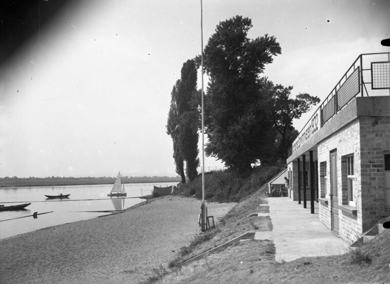 Das Bild zeigt das Hochheimer Strandbad am Ufer des Mains mit einem modernen Gebäude und einer Terrasse im Vordergrund. Am Sandstrand sind einige Boote und ein Segelboot auf dem Wasser zu sehen. Große Bäume säumen das Ufer und sorgen für eine ruhige und idyllische Atmosphäre.