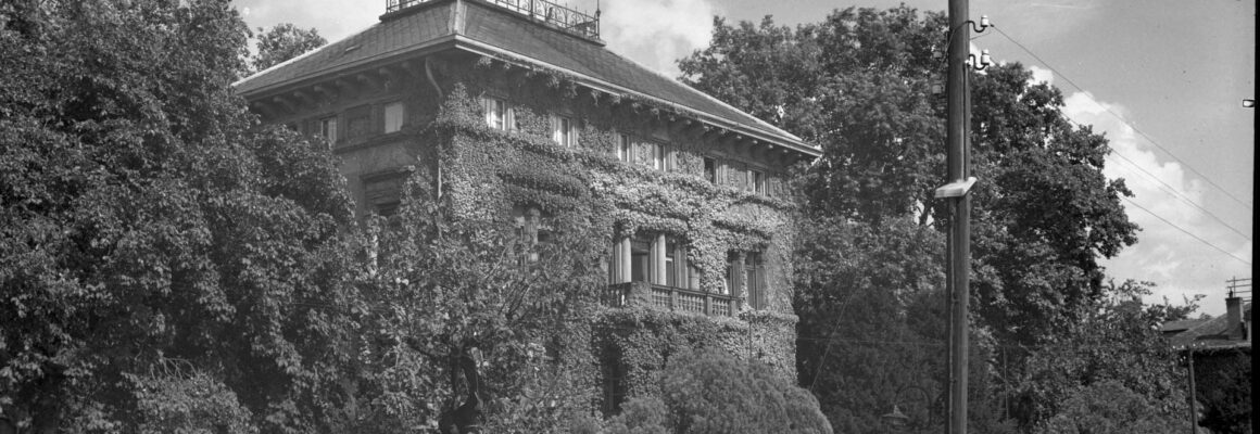 Das Bild in Schwarzweiß zeigt die mit Efeu bewachsene Villa Burgeff, die hinter einer steinernen Mauer und einem schmiedeeisernen Zaun liegt. Das Gebäude hat ein großes, flaches Dach mit einer dekorativen Balustrade und ist von üppigen Bäumen und Büschen umgeben.