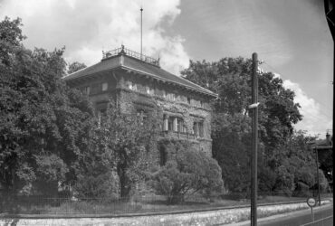 Das Bild in Schwarzweiß zeigt die mit Efeu bewachsene Villa Burgeff, die hinter einer steinernen Mauer und einem schmiedeeisernen Zaun liegt. Das Gebäude hat ein großes, flaches Dach mit einer dekorativen Balustrade und ist von üppigen Bäumen und Büschen umgeben.