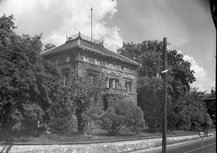 Das Bild in Schwarzweiß zeigt die mit Efeu bewachsene Villa Burgeff, die hinter einer steinernen Mauer und einem schmiedeeisernen Zaun liegt. Das Gebäude hat ein großes, flaches Dach mit einer dekorativen Balustrade und ist von üppigen Bäumen und Büschen umgeben.