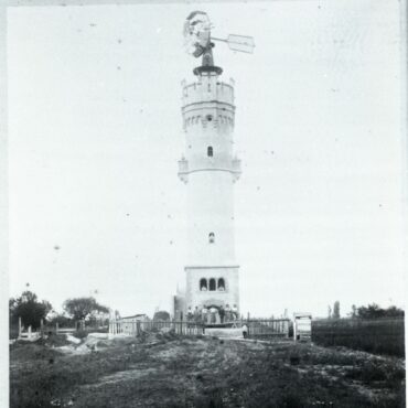 Das Bild zeigt einen Wasserturm mit einem Windrad an der Spitze, umgeben von einem einfachen Zaun. Vor dem Turm sind mehrere Menschen zu sehen, die sich vor dem Eingang versammelt haben, was die Größe und Bedeutung des Bauwerks in der Landschaft betont.