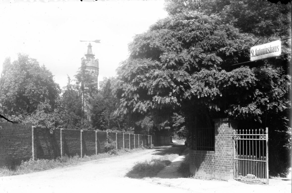 Das Schwarz-Weiß-Foto zeigt den Wasserturm der Firma Burgeff im Park des St. Antoniushauses, umgeben von dichten Bäumen. Im Vordergrund verläuft ein unbefestigter Weg entlang einer überwucherten Ziegelmauer. Rechts ist ein offenes schmiedeeisernes Tor mit einem Schild "St. Antoniushaus" sichtbar.