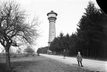 Das Foto zeigt einen hohen Wasserturm, der entlang einer von Bäumen gesäumten Straße steht. Zwei Kinder posieren vor dem Turm, der von einem dichten Wald umgeben ist, was auf eine ländliche oder waldreiche Umgebung hindeutet.