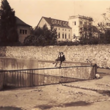 Das Bild zeigt zwei Personen, die auf einem Geländer der Hochheimer Backeswied sitzen. Im Hintergrund ist eine Steinmauer und eine Villa mit der Aufschrift "GRAEGER" zu sehen. Die Szene wirkt ruhig und lässt auf entspannte Atmosphäre schließen.