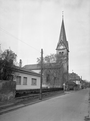 Das Foto zeigt die evangelische Kirche in Hochheim, die mit ihrem markanten, hohen Kirchturm das Straßenbild prägt. Der Turm ist mit einer Uhr versehen und von einem spitzen Dach abgeschlossen, das von einer Wetterfahne gekrönt wird. Entlang der ruhigen Straße, an der die Kirche liegt, befinden sich weitere Gebäude.