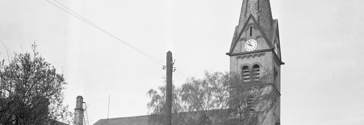 Das Bild zeigt den Turm der evangelischen Kirche in Hochheim mit einer spitzen, schiefergedeckten Turmspitze und einer großen Uhr an der Vorderseite.