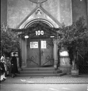 Der Eingang einer evangelischen Kirche, geschmückt mit Girlanden und einem großen Schild mit der Zahl „100“. Eine kleine Gruppe von Menschen, darunter Kinder, steht vor dem Eingang und betrachtet das Geschehen.