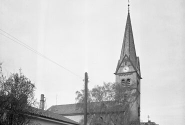 Das Bild zeigt den Turm der evangelischen Kirche in Hochheim mit einer spitzen, schiefergedeckten Turmspitze und einer großen Uhr an der Vorderseite.