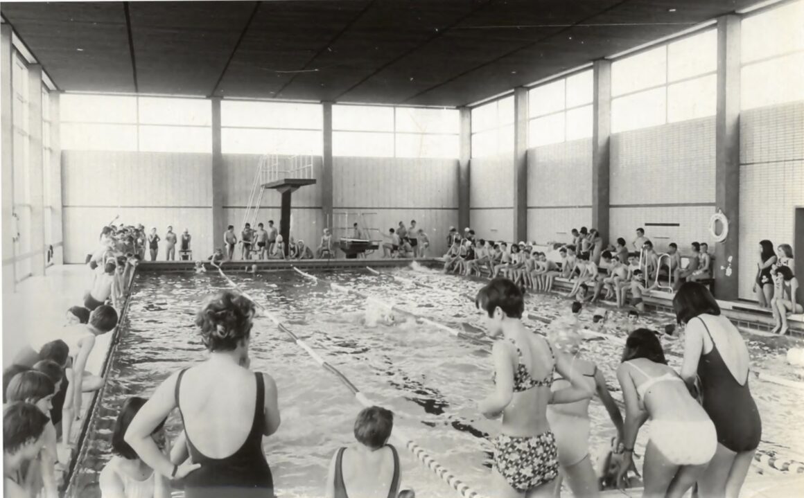 Eine große Gruppe von Kindern und Jugendlichen versammelt sich in einem Hallenbad für ein Schwimmereignis. Am Beckenrand stehen und sitzen viele Zuschauer, während einige Schwimmer im Wasser an einem Wettkampf teilnehmen. Die Stimmung ist lebhaft, und das Schwimmbad ist gut gefüllt.