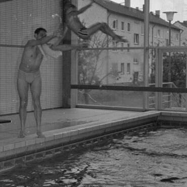 Eine Person wirft ein Kind spielerisch ins Schwimmbecken. Beide scheinen Freude zu haben. Im Hintergrund sind Fenster, durch die man Wohngebäude außerhalb des Schwimmbades sehen kann.