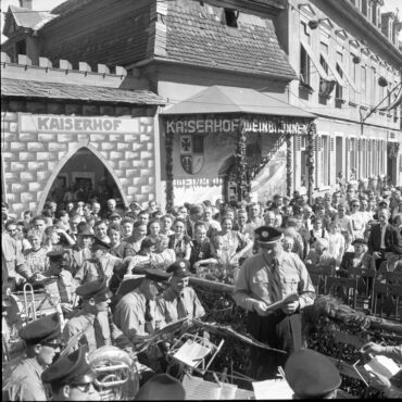 Eine große Menschenmenge hat sich zum Weinfest 1950 vor dem Kaiserhof versammelt. Im Vordergrund spielt eine Blaskapelle und ein Mann in Uniform liest von einem Blatt Papier vor. Im Hintergrund sind weitere Zuschauer zu sehen, die das Geschehen verfolgen.