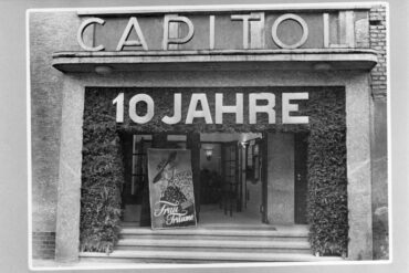 Das Bild zeigt den Eingang des Capitol-Kinos, das mit einer großen "10 Jahre" Dekoration geschmückt ist, um sein zehnjähriges Jubiläum zu feiern. Vor dem Eingang steht ein Plakat des Films "Frau Träume" mit Marika Rökk. Die Fassade ist mit Laubwerk dekoriert, und durch die offenen Türen kann man das Innere des Kinos erkennen.