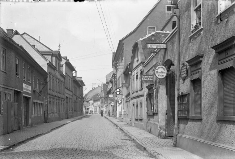 Das Foto zeigt eine historische Straßenszene in Hochheim. Auf der rechten Seite des Bildes sind mehrere Geschäfte und Gastwirtschaften zu sehen, darunter das Lichtspielhaus, das Weine und Speisen anbietet. Die gepflasterte Straße wirkt ruhig, und nur wenige Passanten sind in der Ferne zu erkennen.