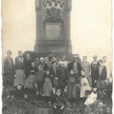 Eine Gruppe von etwa 20 Menschen, darunter Männer, Frauen und Kinder, posiert vor dem Königin-Victoria-Denkmal in Hochheim. Die Personen sind schlicht gekleidet, und das Bild wirkt durch seinen vergilbten Zustand sehr alt.