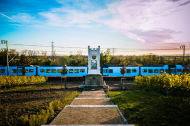 Das Bild zeigt das Königin-Victoria-Denkmal in Hochheim, im Vordergrund von Weinreben umgeben. Im Hintergrund fährt ein blauer Zug vorbei, der eine dynamische Kulisse vor dem ruhig stehenden, historischen Denkmal bietet. Die Szene vereint Geschichte und Moderne in einer idyllischen Landschaft.