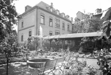 Das Foto zeigt den Garten des Krankenhauses mit einem Brunnen und Parkbänken vor einem Backsteingebäude. Der Garten ist üppig bepflanzt und bietet einen ruhigen Rückzugsort für Patienten und Besucher.