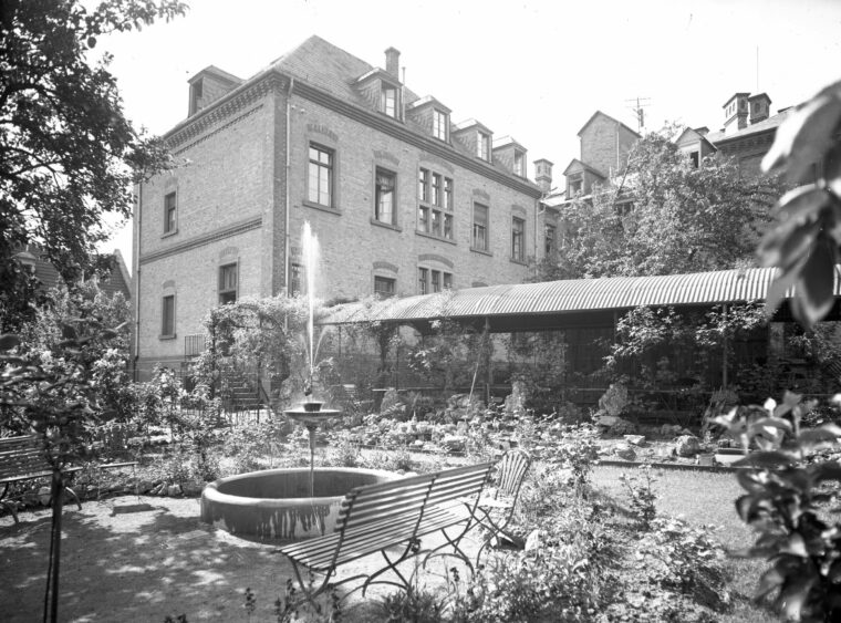 Das Foto zeigt den Garten des Krankenhauses mit einem Brunnen und Parkbänken vor einem Backsteingebäude. Der Garten ist üppig bepflanzt und bietet einen ruhigen Rückzugsort für Patienten und Besucher.