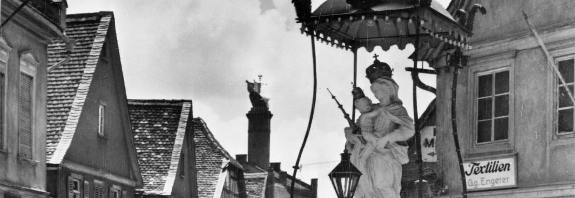 Die Madonna mit Kind steht in der Mitte eines alten Marktplatzes, umgeben von Fachwerkhäusern. Der Himmel ist leicht bewölkt und die Atmosphäre wirkt ruhig.