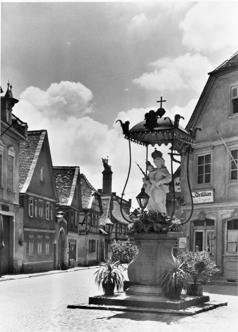 Die Madonna mit Kind steht in der Mitte eines alten Marktplatzes, umgeben von Fachwerkhäusern. Der Himmel ist leicht bewölkt und die Atmosphäre wirkt ruhig.